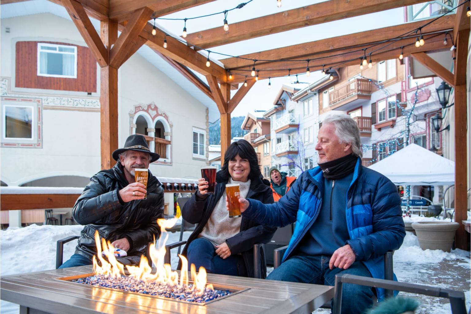 Three people toasting drinks by an outdoor fire in a snowy village setting.
