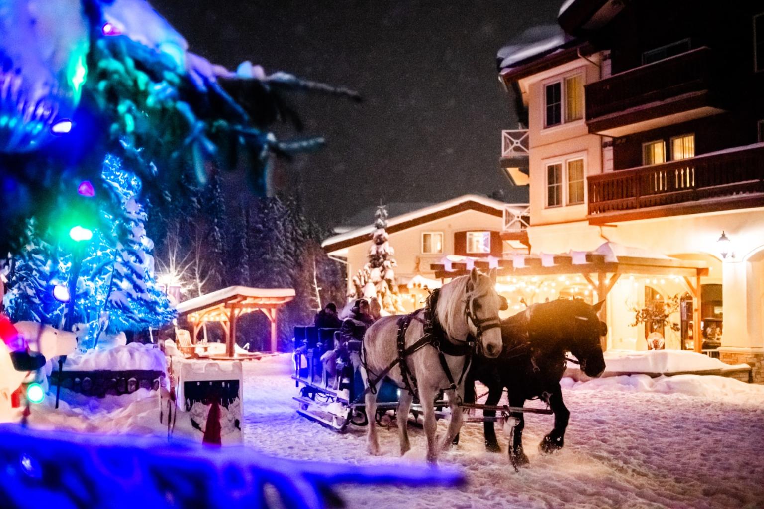Horse-drawn sleigh in snowy village, festive lights glow warmly.