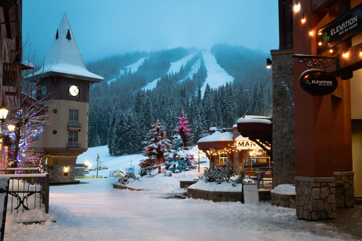 Snowy village street with lights, clock tower, and snowy mountains in the background.