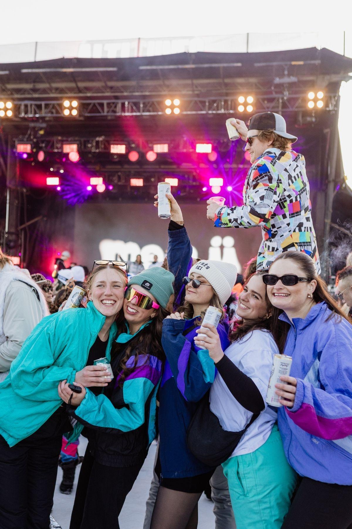 Festival crowd in colorful jackets and sunglasses, holding drinks, with a lit stage in the background.