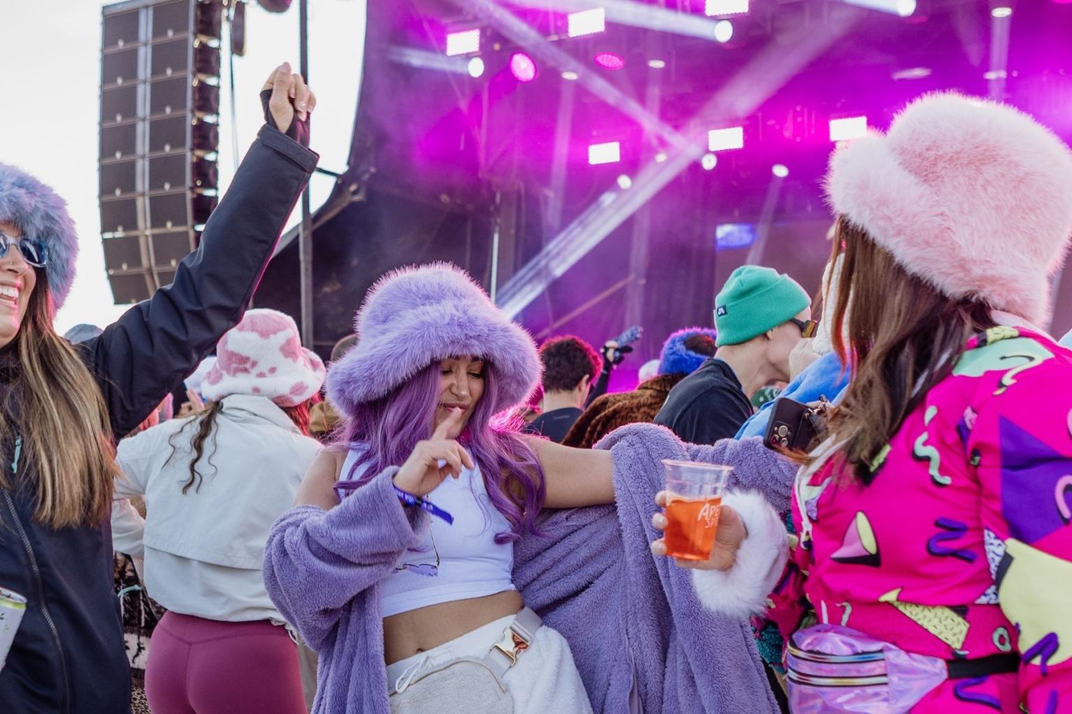 Festival crowd in colorful outfits, purple lighting, people dancing and enjoying music.