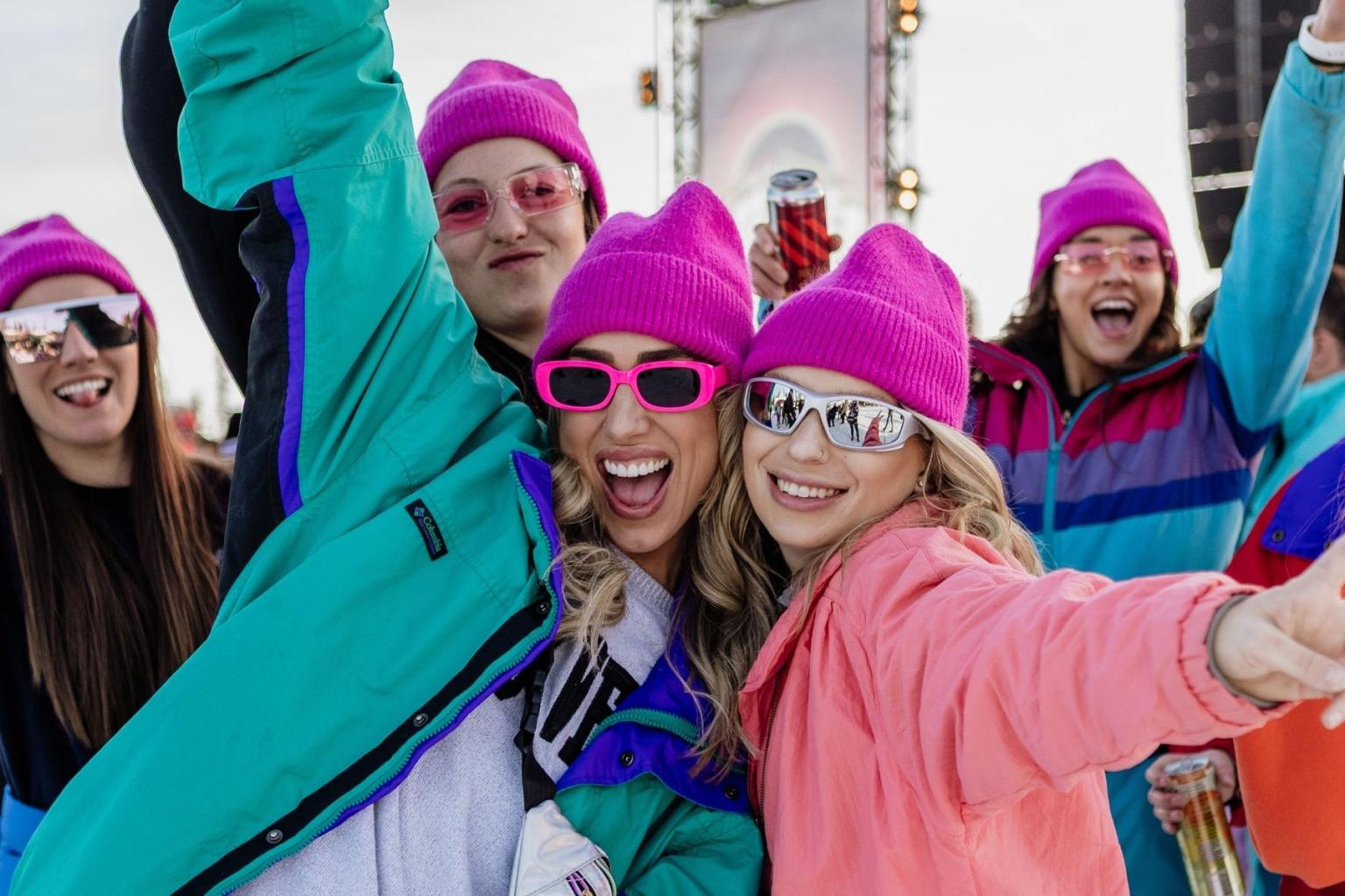 Friends in colorful winter clothes and pink beanies, smiling and posing outdoors.