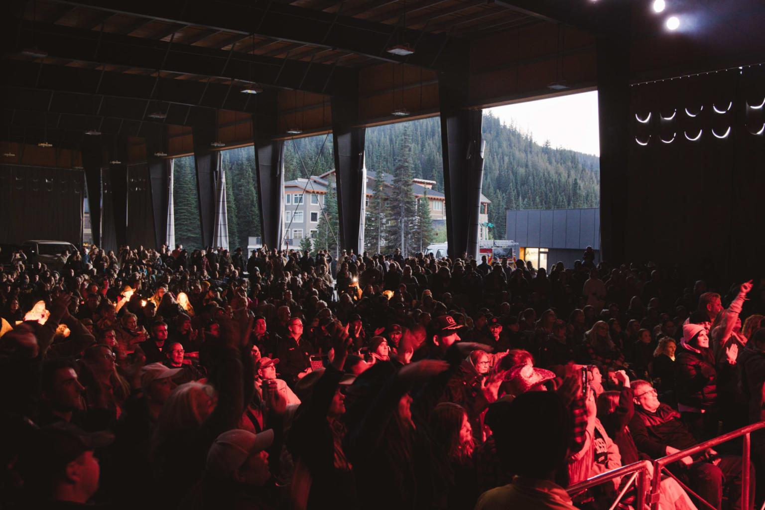 Concert crowd under red lighting in large venue with forest view.
