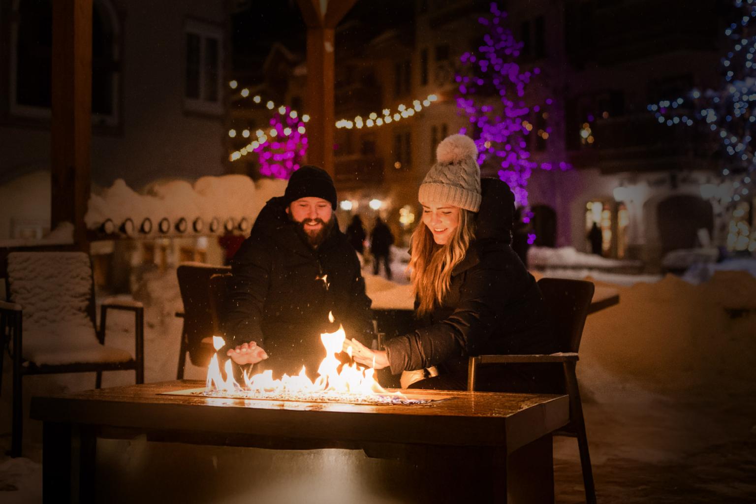 Two people smiling around an outdoor fire pit in a snowy, illuminated setting.