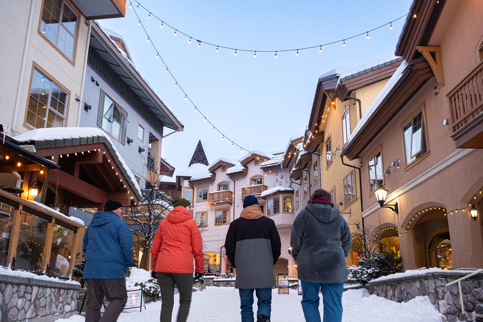 Four people walk down a snowy village street with string lights above.