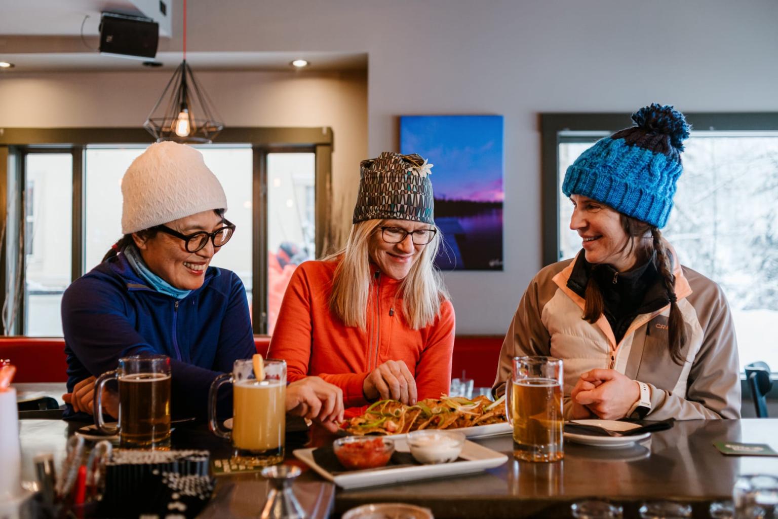 Three friends in winter hats enjoy drinks and food at a cozy bar.