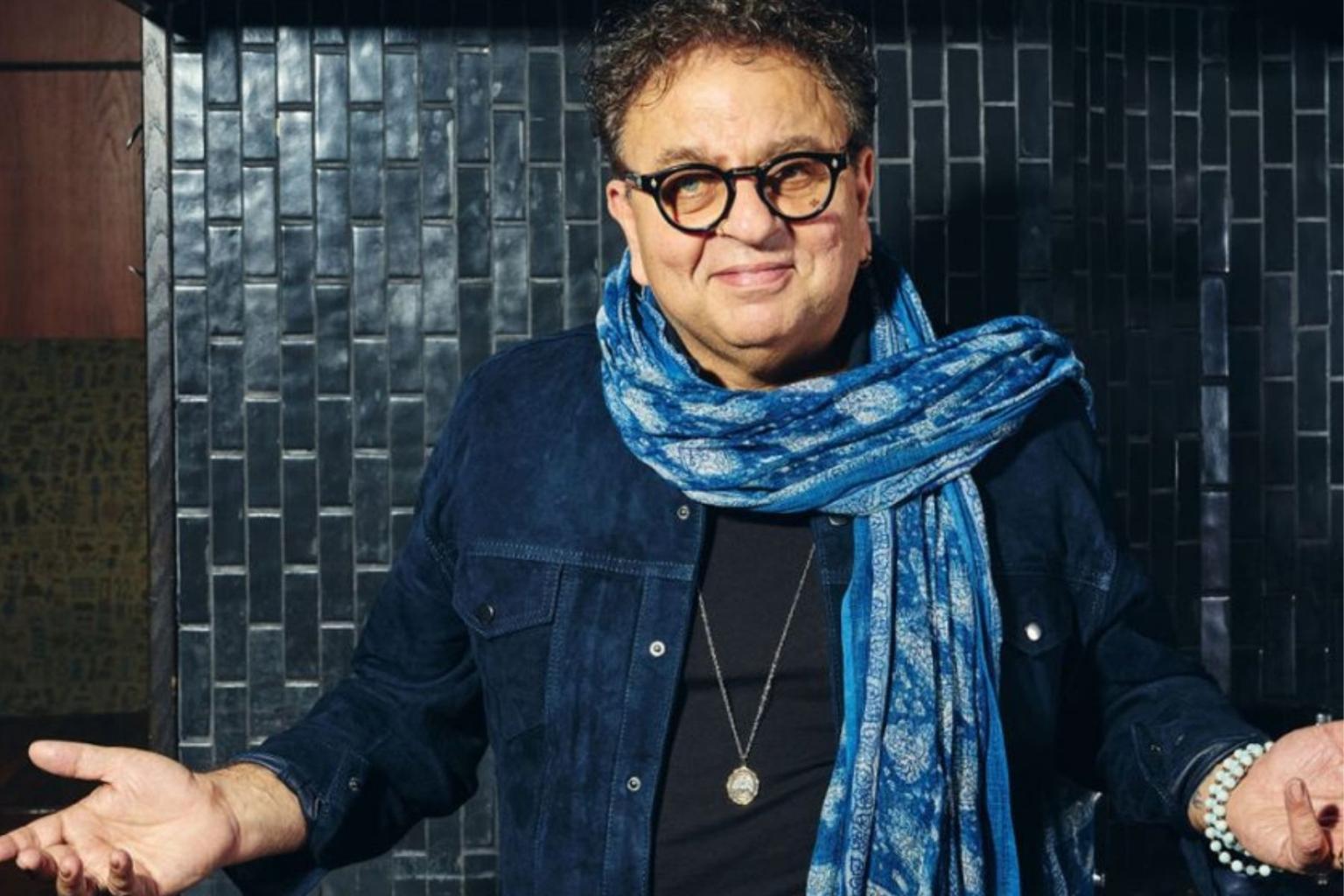 Vikram Vij smiling with outstretched arms, wearing a blue scarf and glasses.