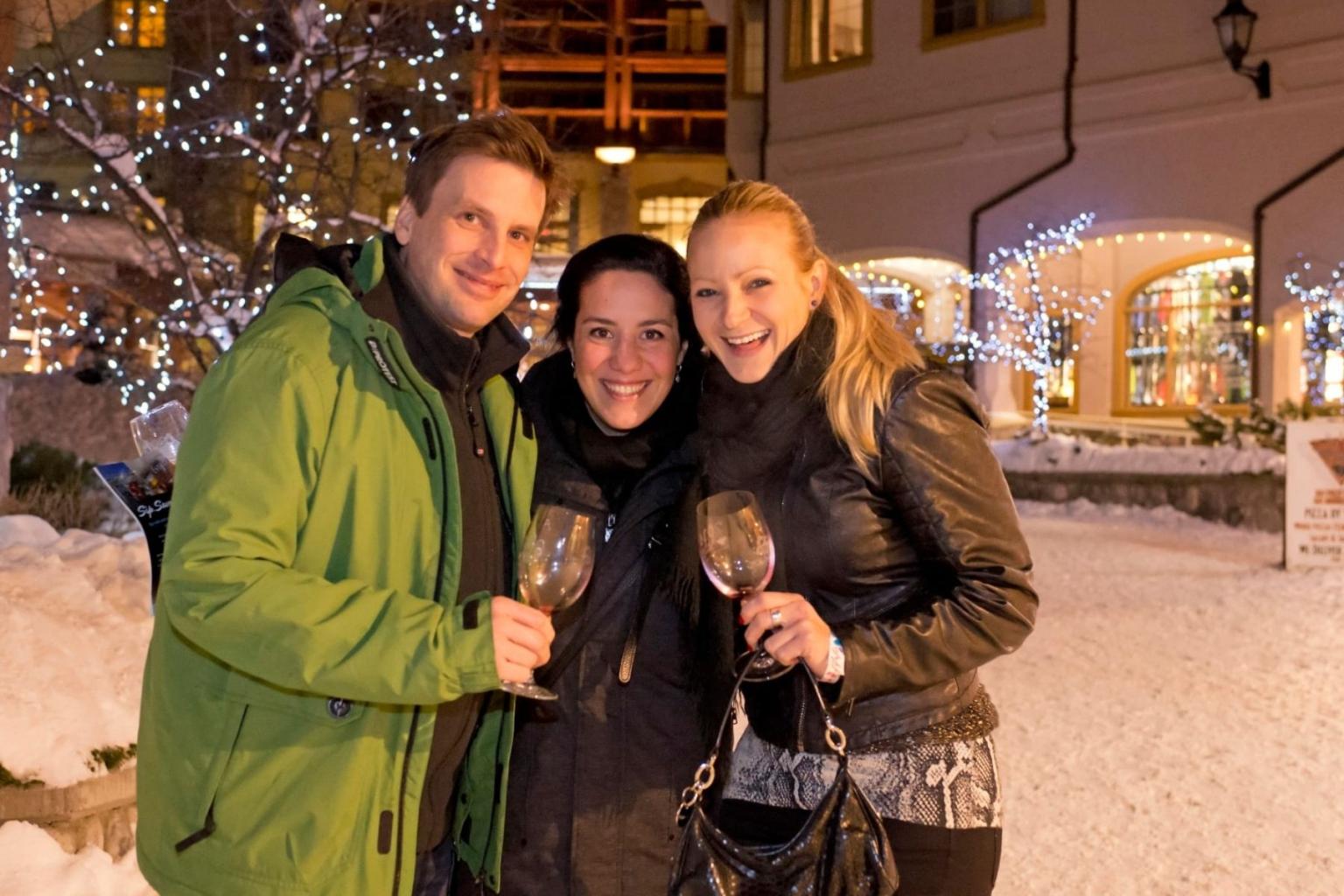 Three people smiling with wine glasses, standing in a snowy, festive setting.