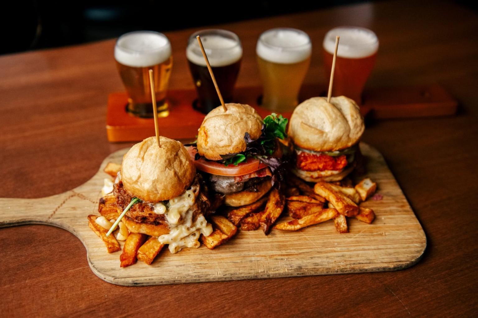 Three sliders with fries on wooden board, beers in the background.