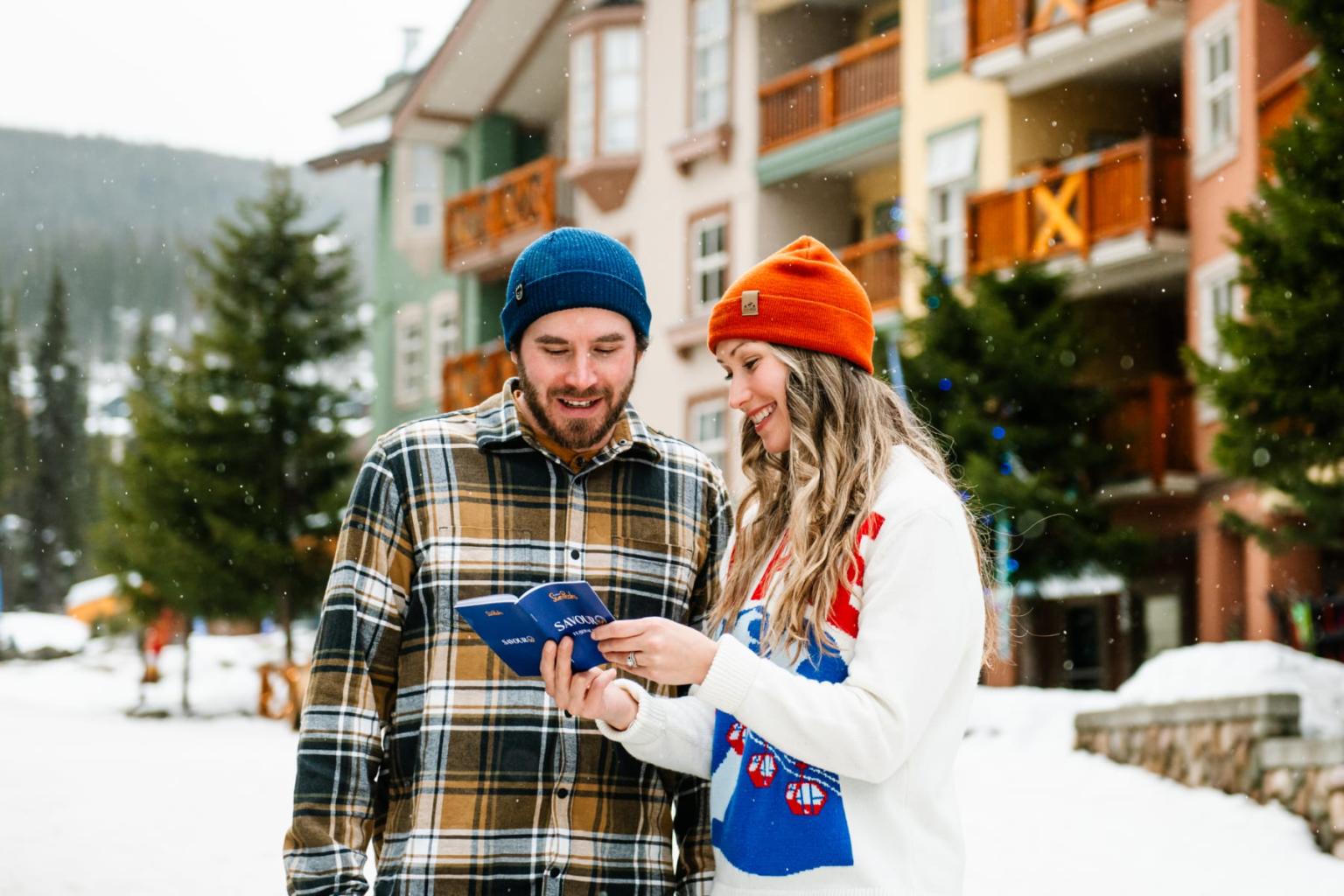 Couple in winter hats looking at a map, standing in a snowy village.