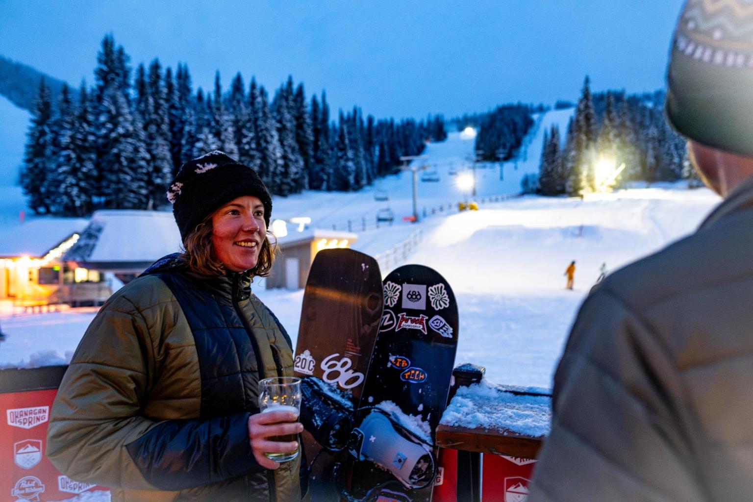 Snowboarders chatting by a snowy ski slope at dusk, under a clear blue sky.
