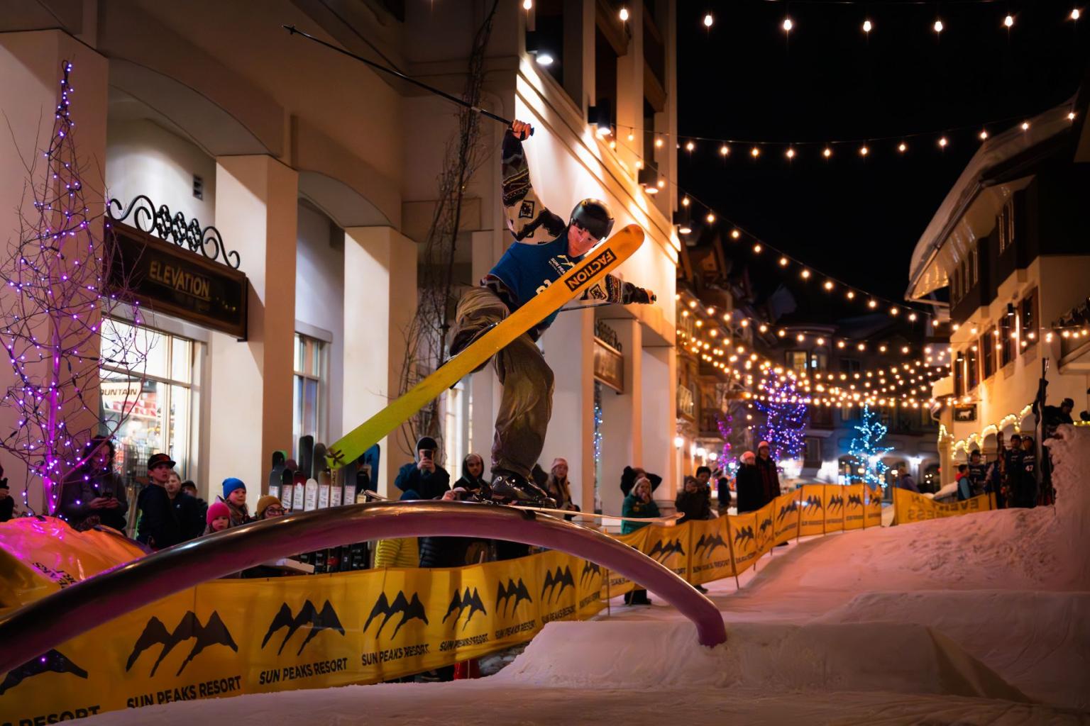 Snowboarder performs a trick on a rail in a night time street setting.