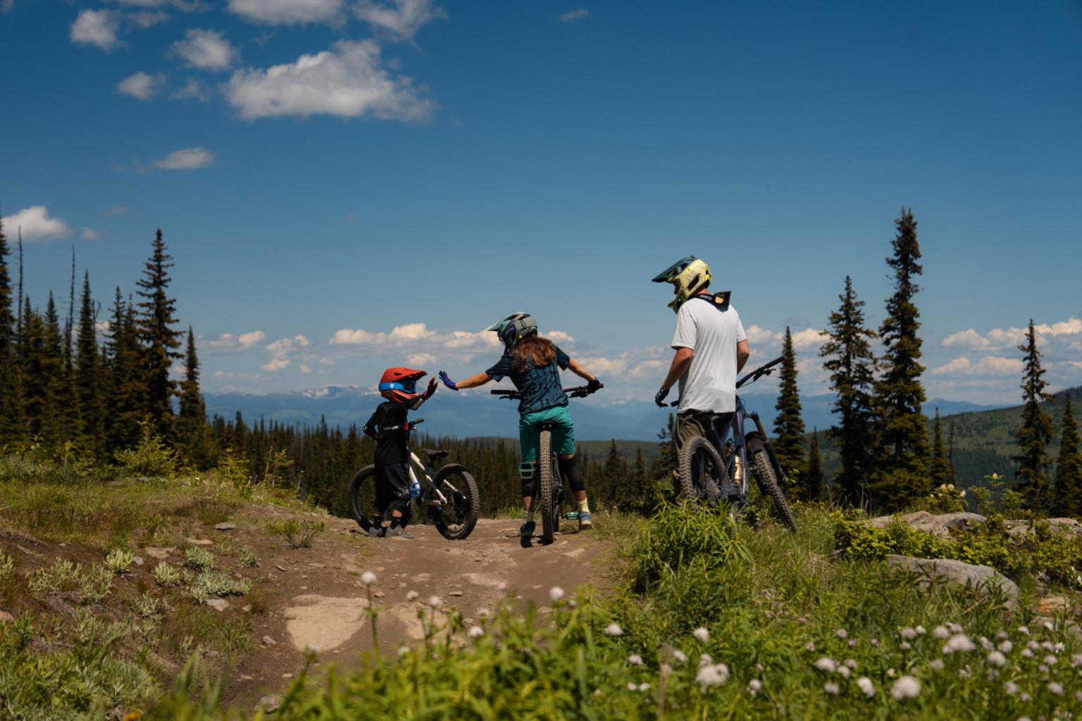 Three cyclists in helmets on a scenic mountain trail under a blue sky.