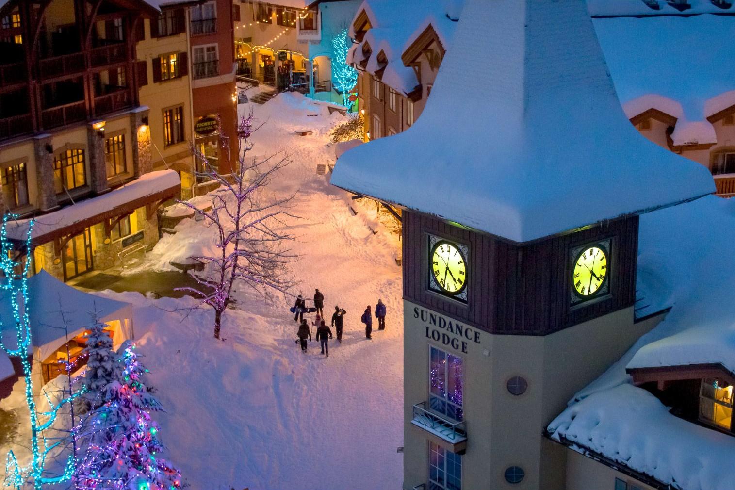 Snow-covered ski village square with lit clock tower at dusk.