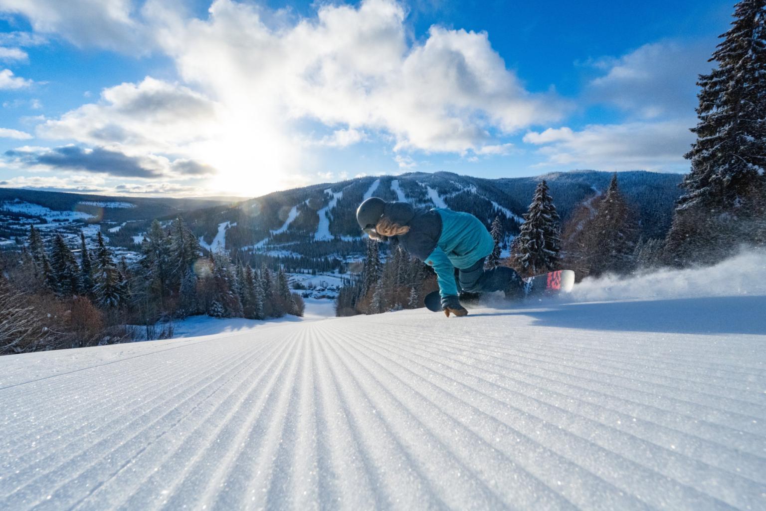 Snowboarder gliding down a sunlit, groomed slope with mountains in the background.