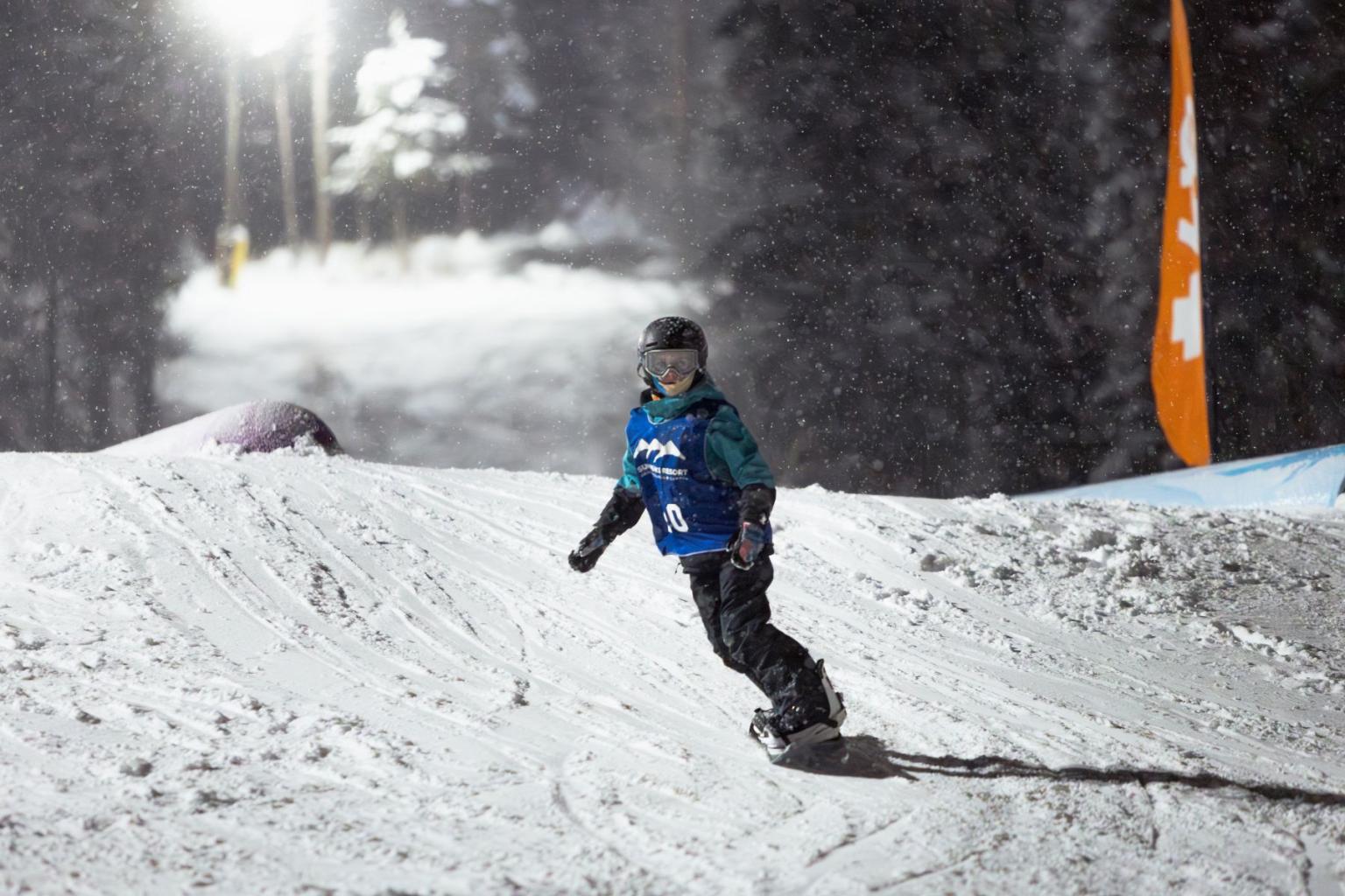 Snowboarder in competition pinny and winter gear, riding on a snowy slope at night.