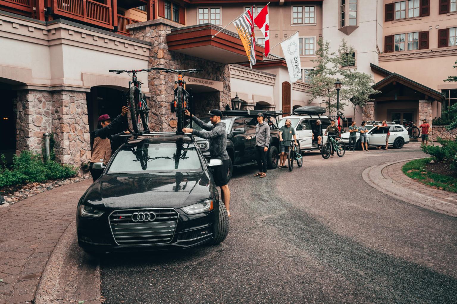 Cars parked in front of a lodge with flags, people loading bikes.