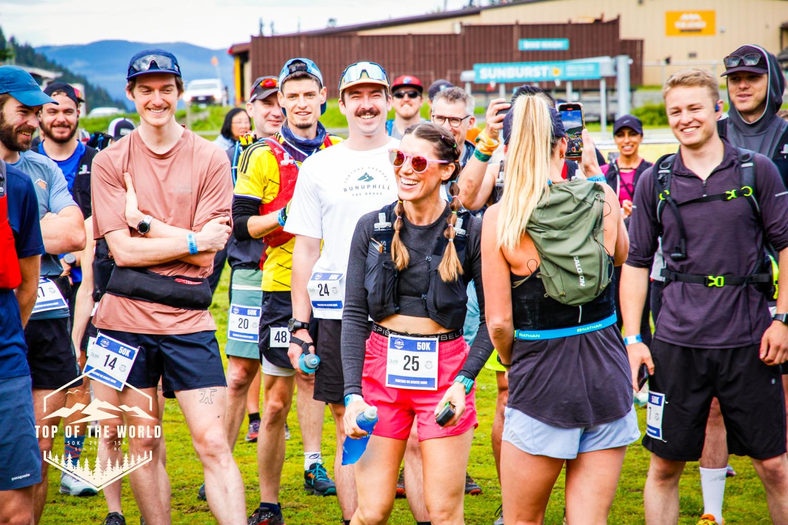 Run Like A Girl event. Runners smiling at a race start line with numbered bibs outdoors. Hexagonal event logo with mountains and text in the bottom left corner, text reads "Top Of The World".