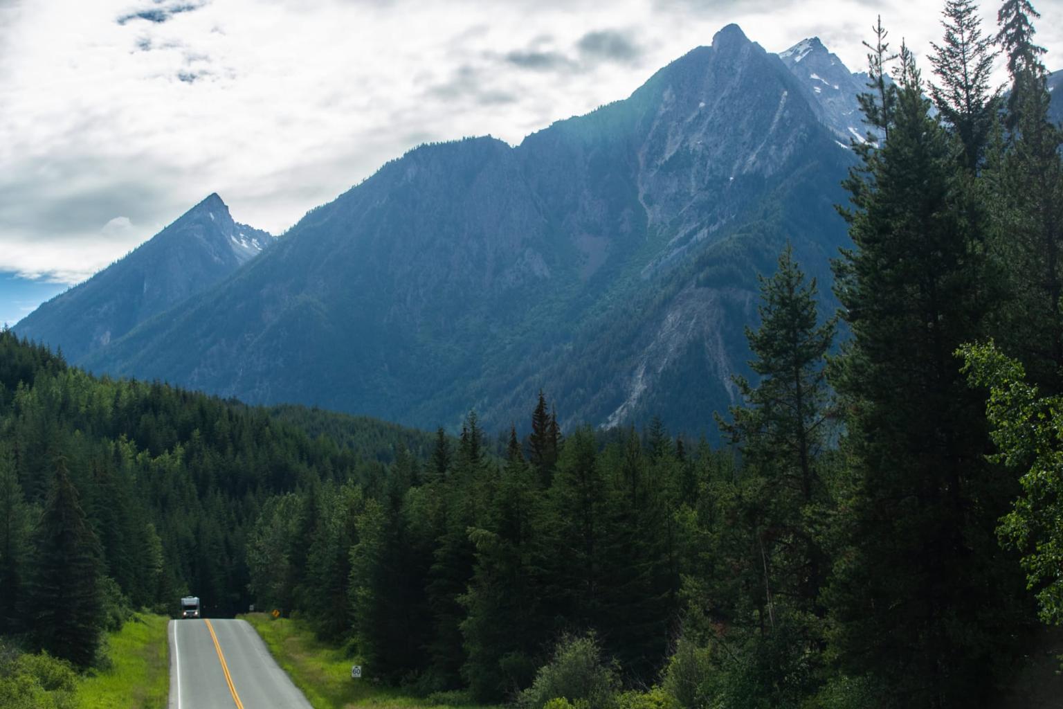 Mountain road lined with dense trees under cloudy sky.
