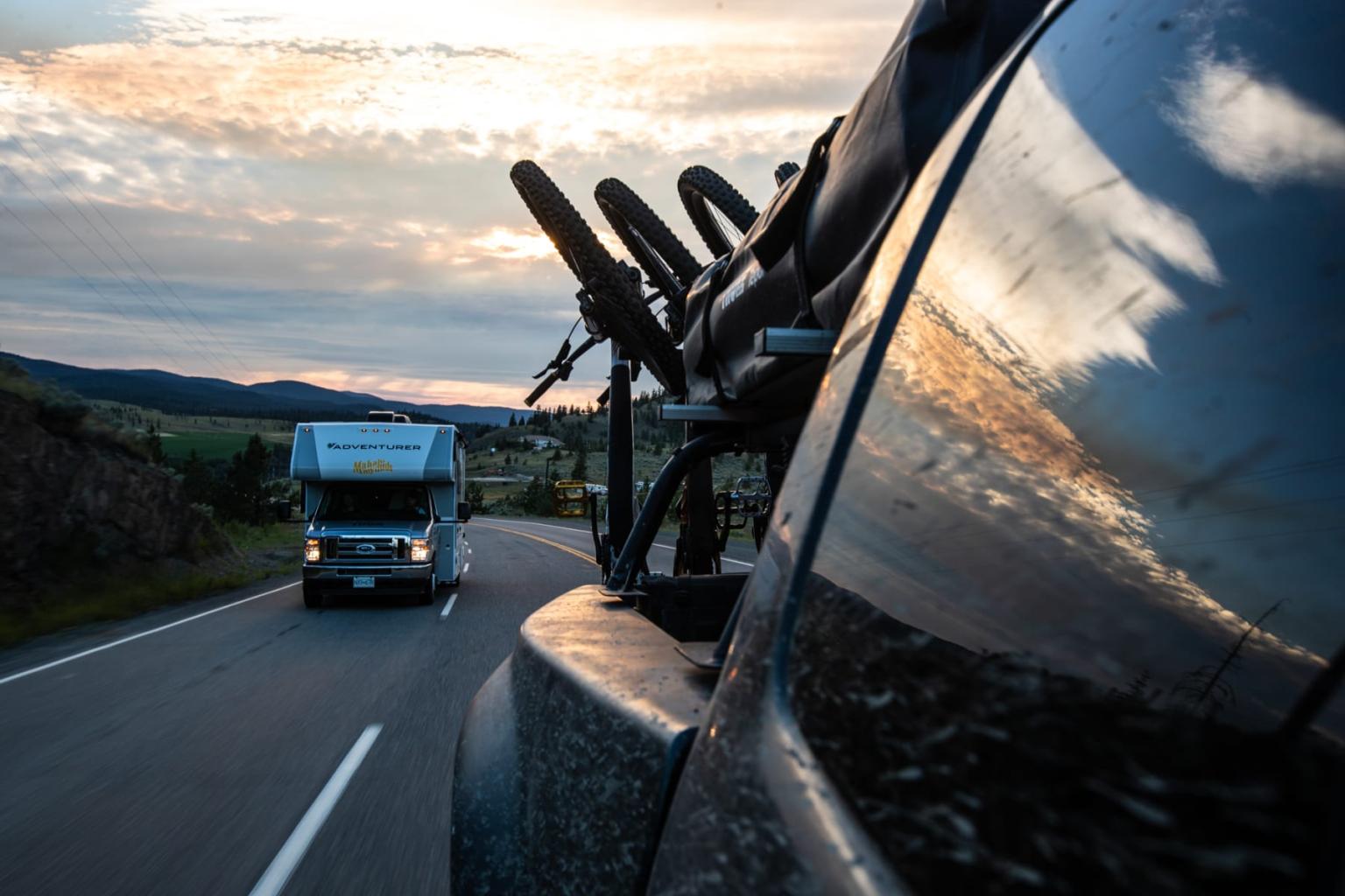Truck and car with bike rack on rural road at sunset.
