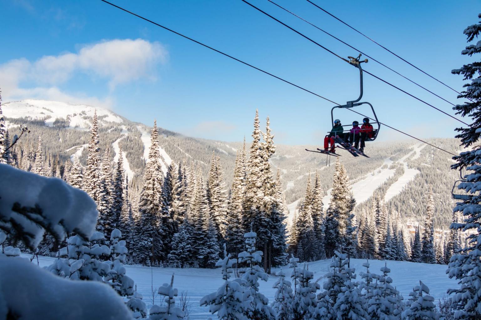 Ski lift with people over snowy trees and mountains, clear blue sky.
