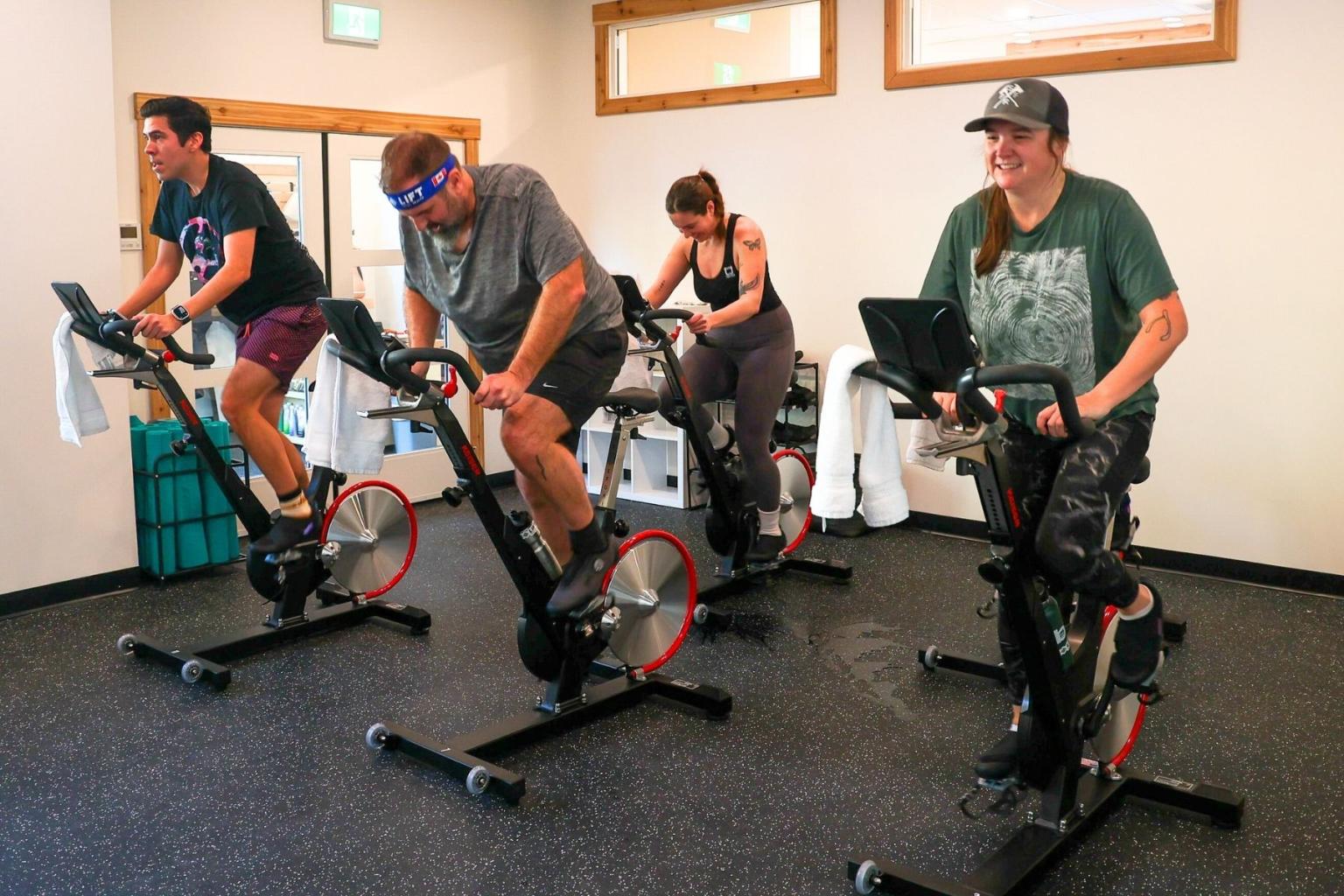Four people exercising on stationary bikes in a gym, appearing focused and energetic.