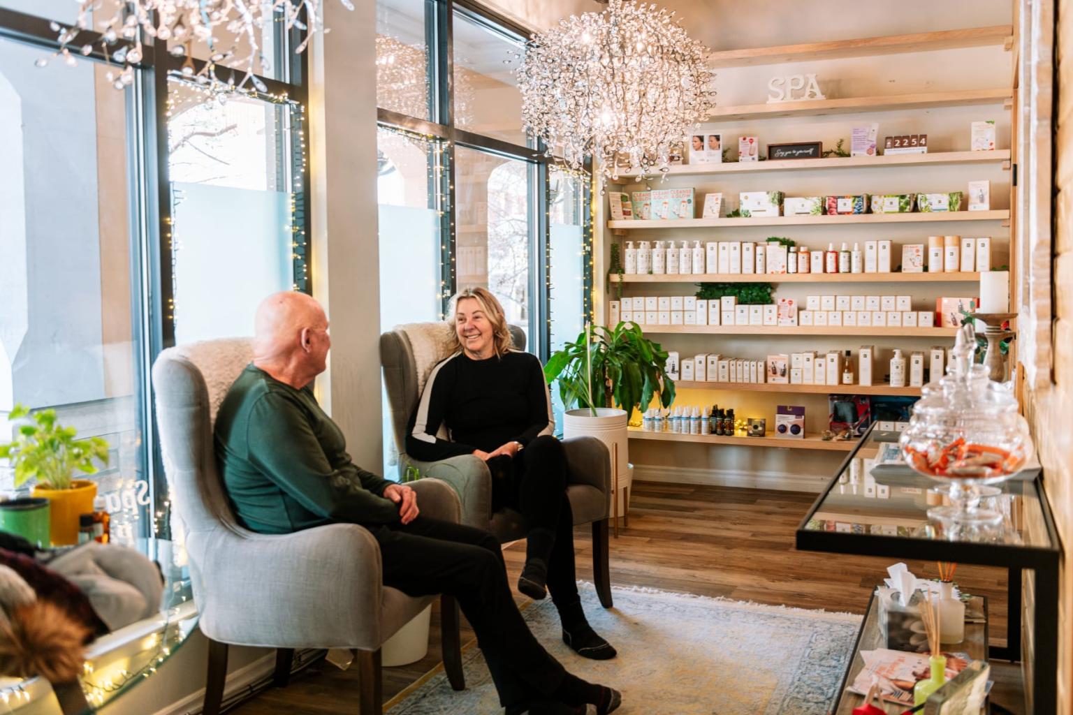 Two people sit in cozy chairs in a warmly lit shop with shelves of products.