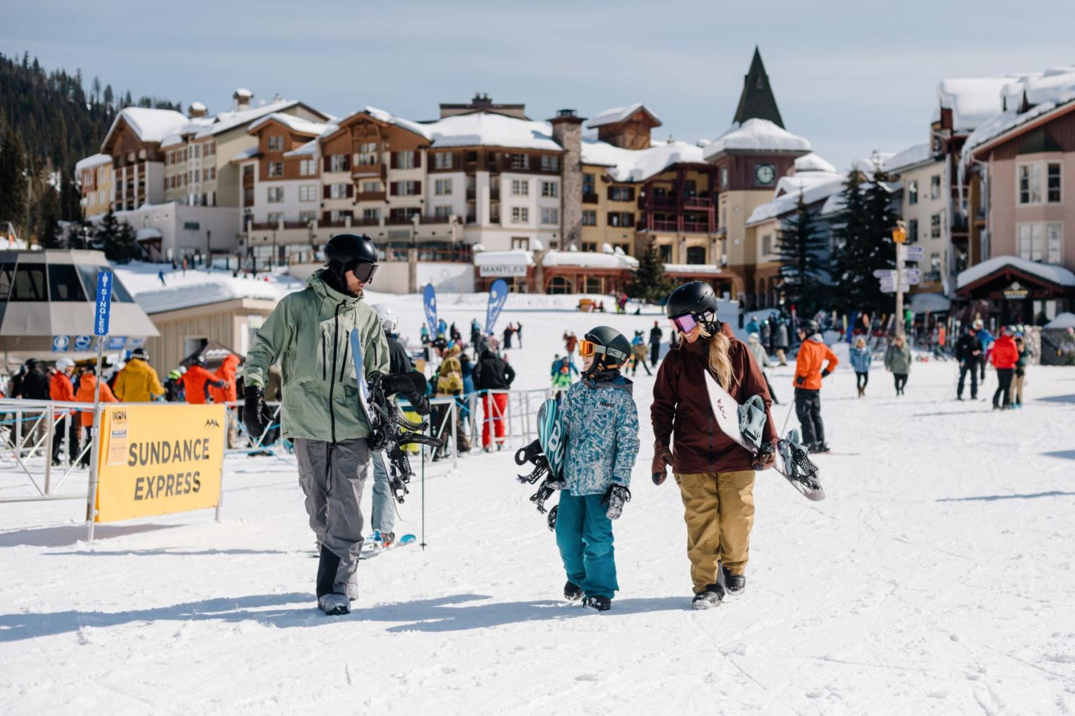 Snowboarder family walking near a snowy resort; buildings in the background.