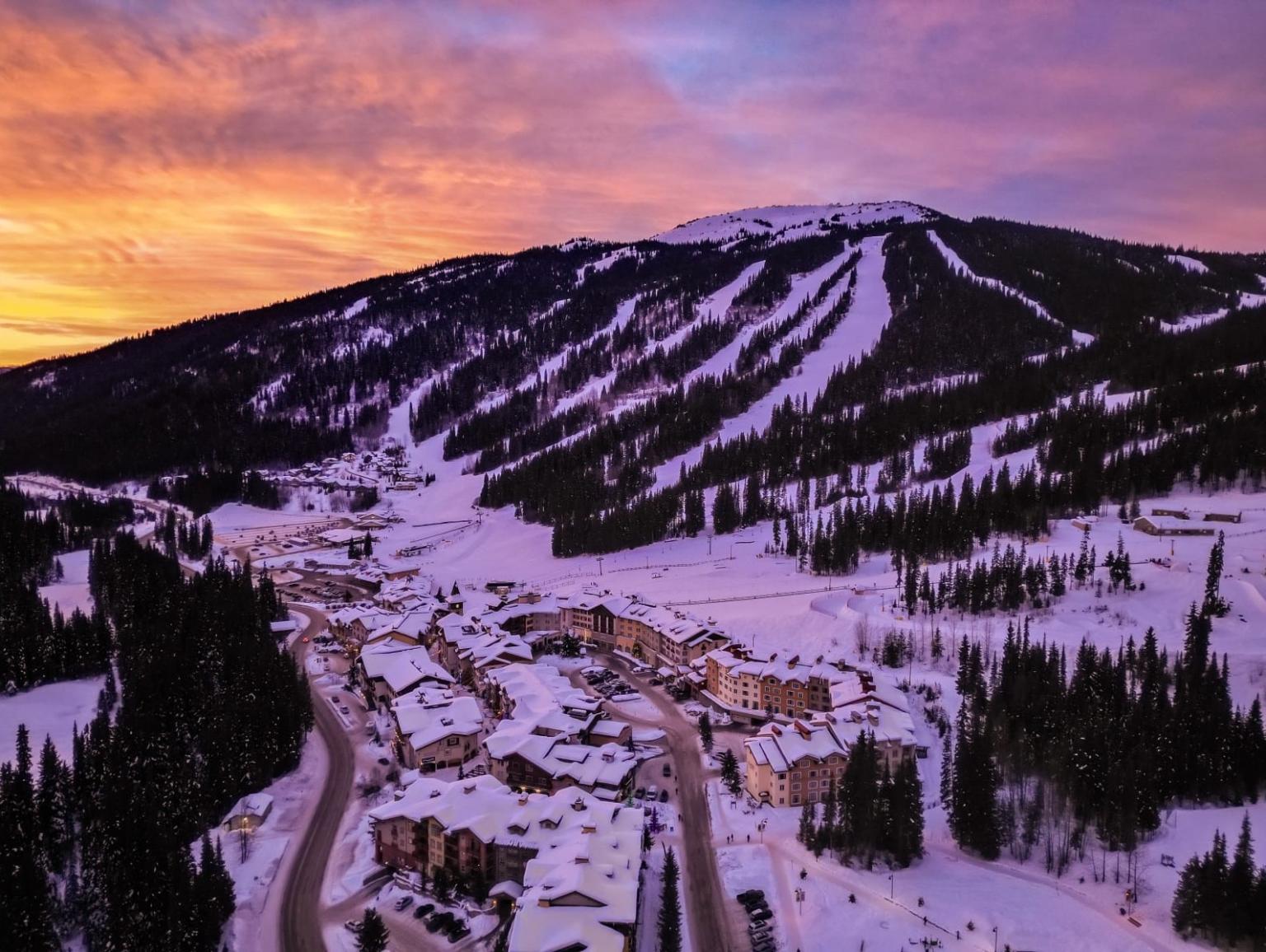 Snowy mountain village at sunset with colorful sky and ski slopes.