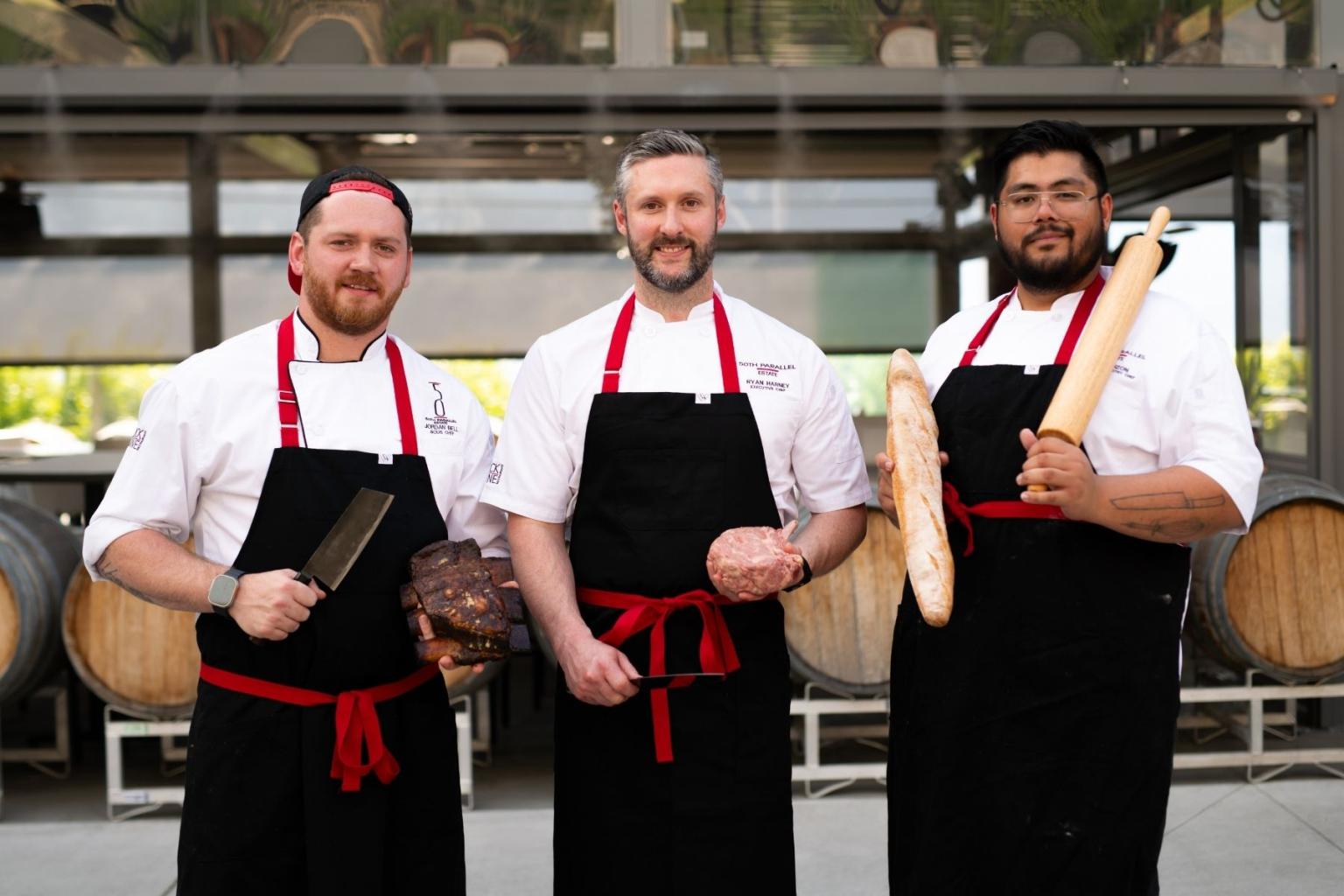 Three chefs in uniforms, each holding kitchen tools, stand in front of wine barrels.