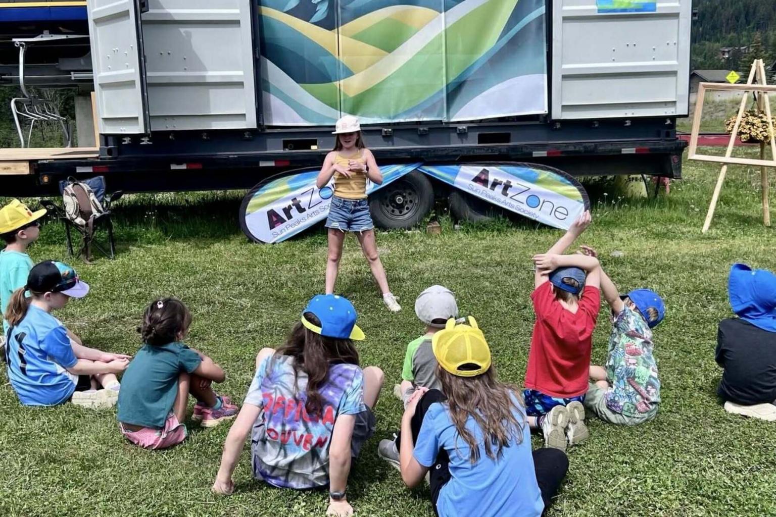 Child performing outdoors for seated group, colorful backdrop, sunny day.