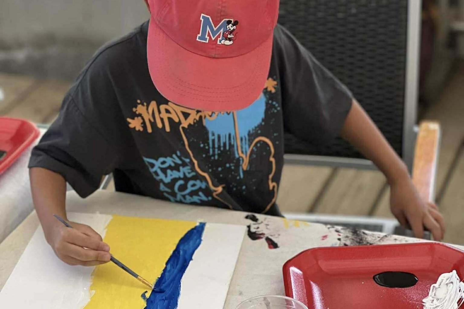 Child painting blue and yellow on paper at a table, wearing a red cap.