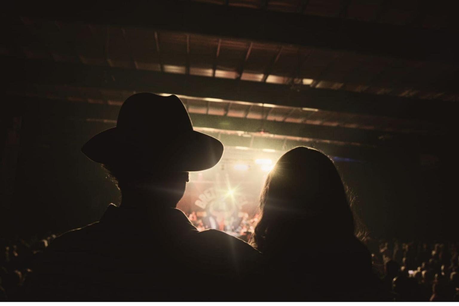 Two silhouettes at a concert, with bright stage lights ahead.
