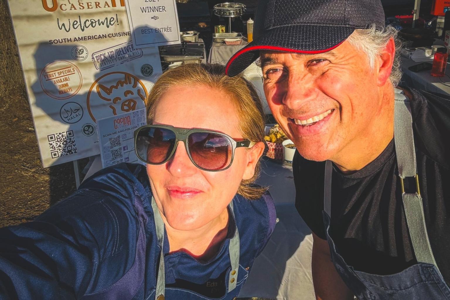 Smiling man and woman in hats and aprons, outdoors in sunlight.