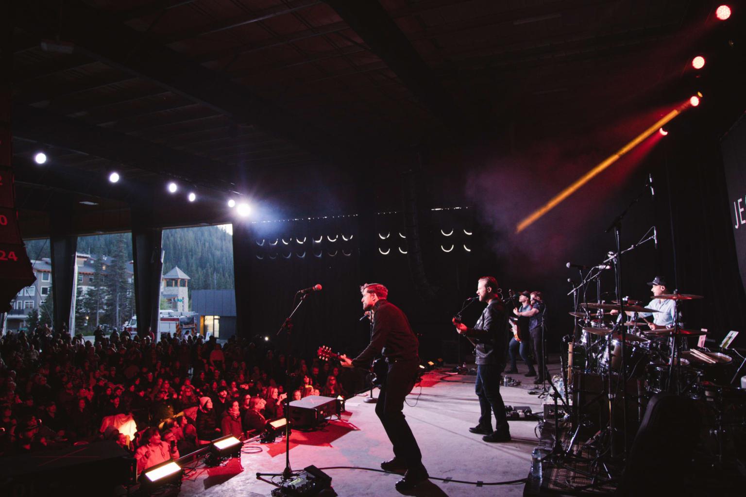 Band performing onstage in a dimly lit concert venue with large audience.