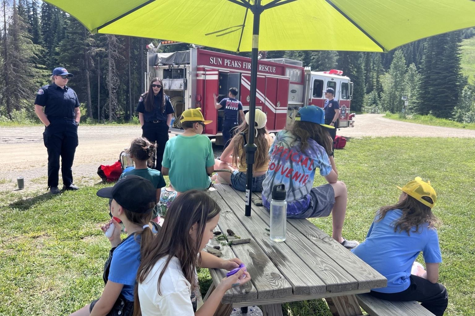 Children at picnic table near a fire truck and firefighters, green umbrella overhead.