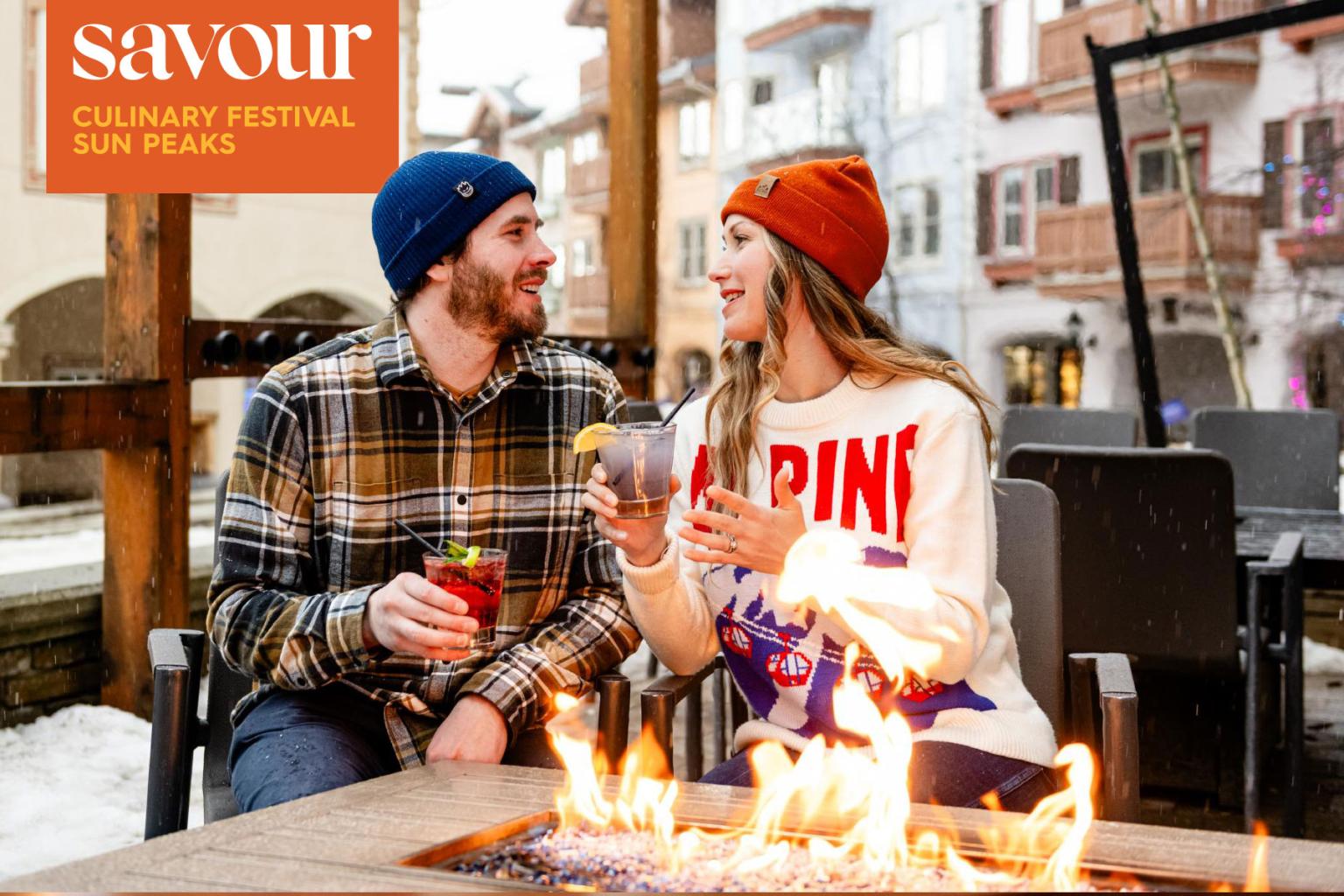 Couple smiling by outdoor fire pit, holding drinks in a snowy town setting.