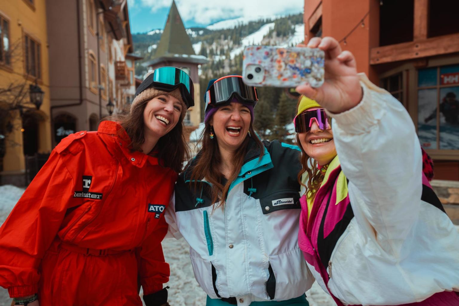 Three friends in colorful ski gear take a selfie in a snowy village.