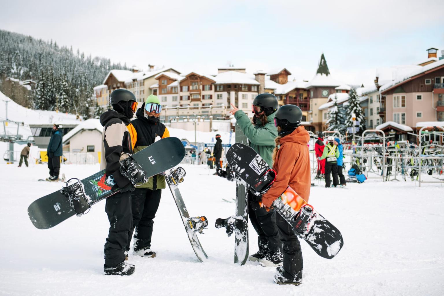 Snowboarders in winter gear chatting on snowy slope, buildings in background.