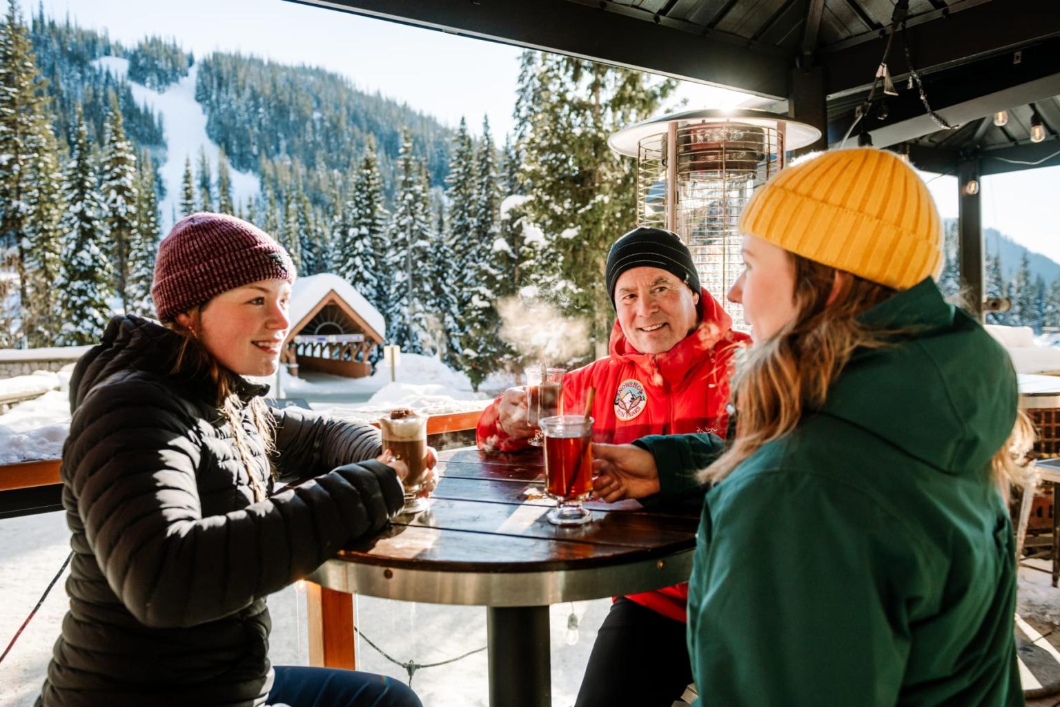 Three people in winter clothes enjoy drinks on a snowy mountain patio.