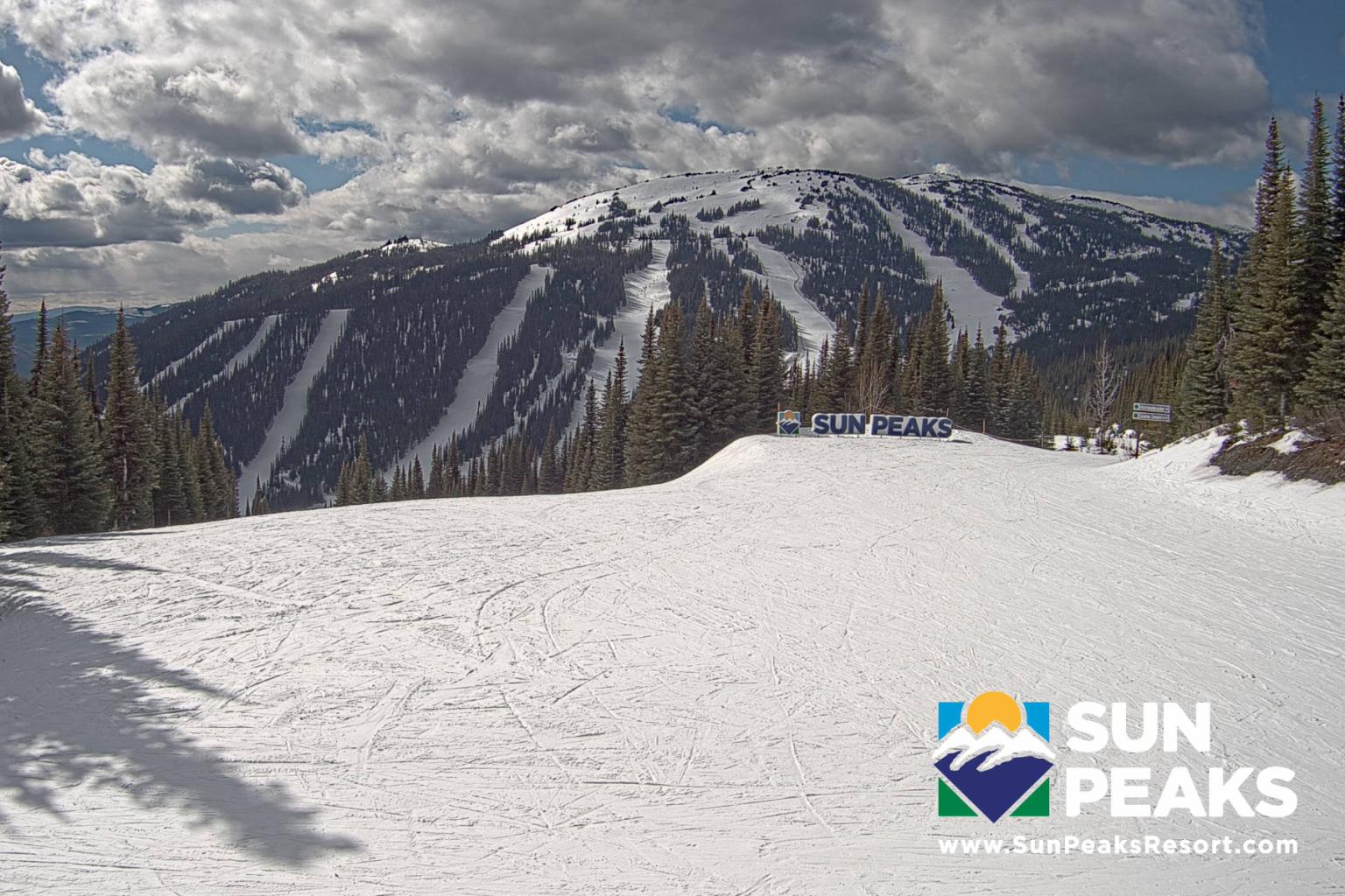 Snow-covered mountain with ski slopes under cloudy sky; Sun Peaks Resort logo in corner.