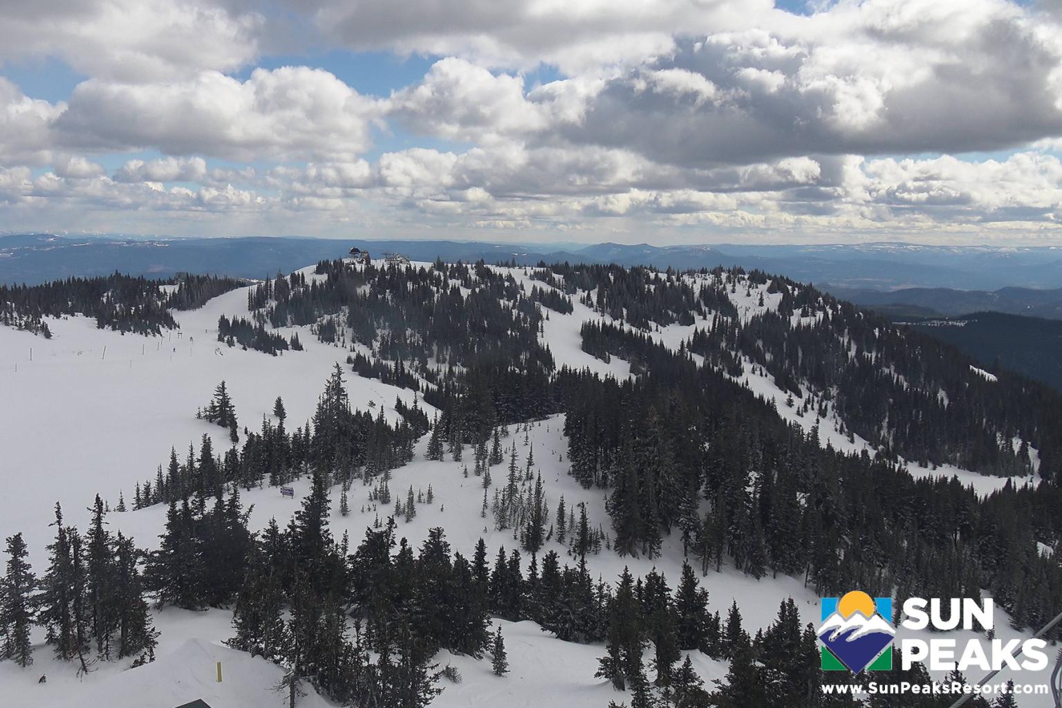 Snow-covered mountain landscape with scattered trees and cloudy sky. Sun Peaks logo in corner.
