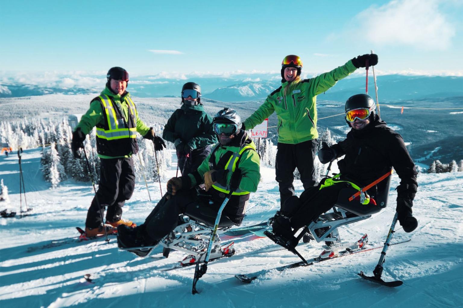 Skiers on a snowy mountain, some seated in adaptive ski equipment, under a clear blue sky.