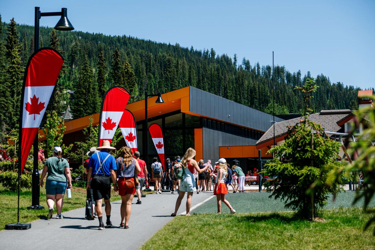 People walking near a building with Canadian flags, surrounded by trees.