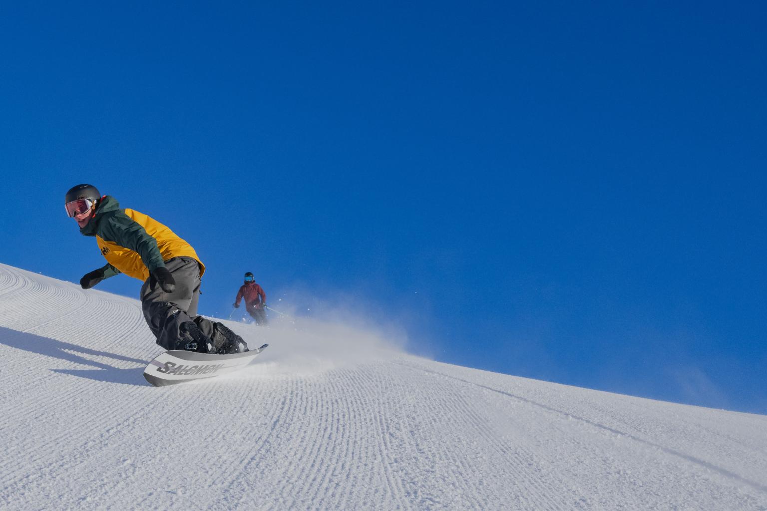 Snowboarder carving down a snowy slope under a clear blue sky.