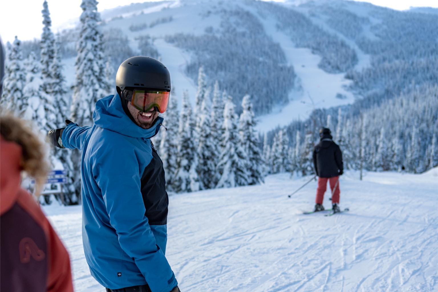 Man smiling on snowy mountain slope, skier in the distance.