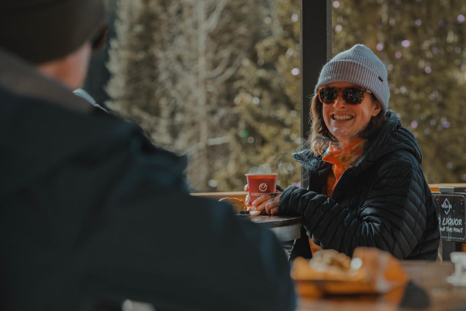 Smiling person in winter clothes holds a coffee cup on a patio near a snowy window.