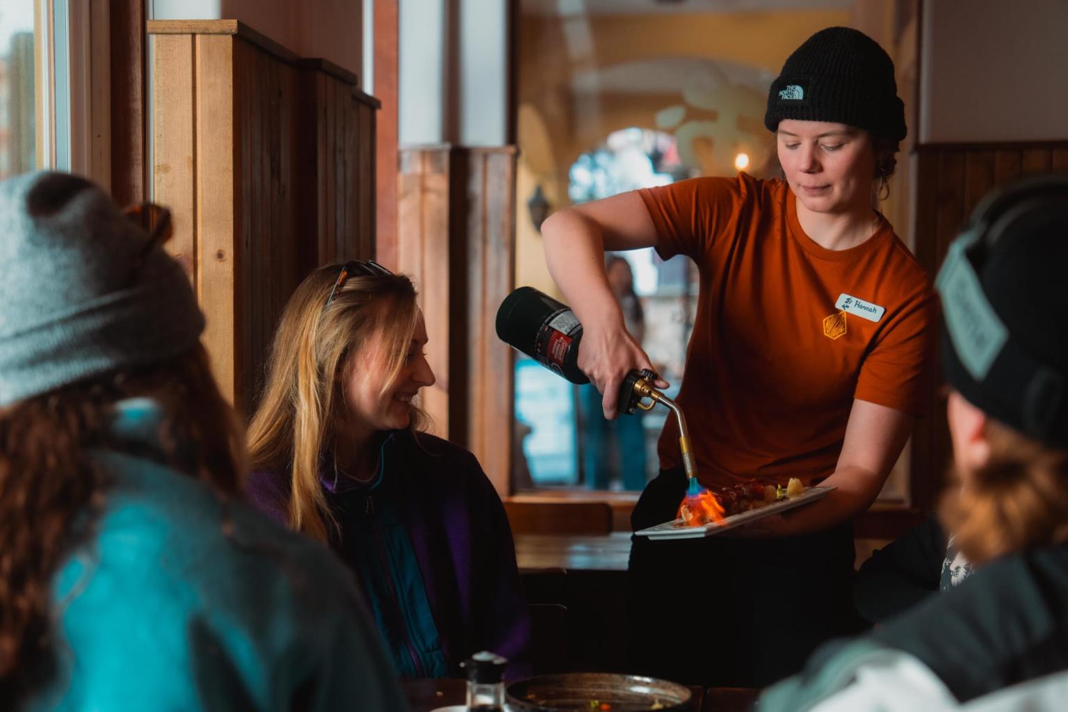 Server flaming oshizushi tableside for smiling woman in a cozy restaurant setting.