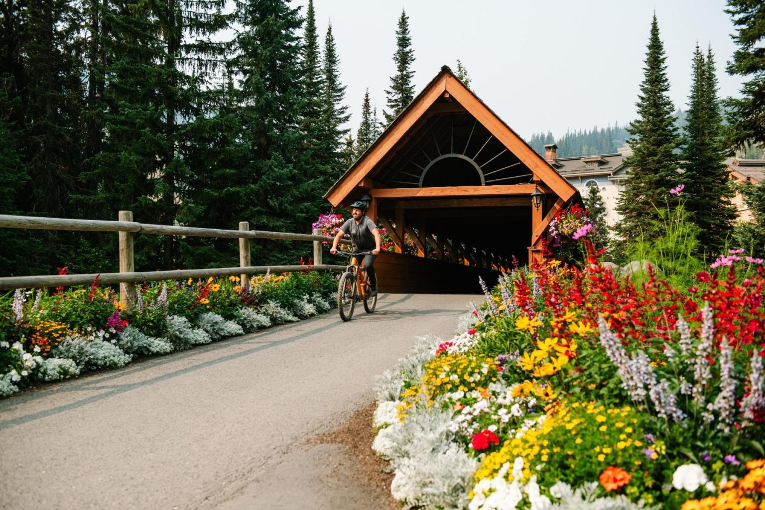 Cyclist on path by flower beds, approaching a wooden covered bridge.