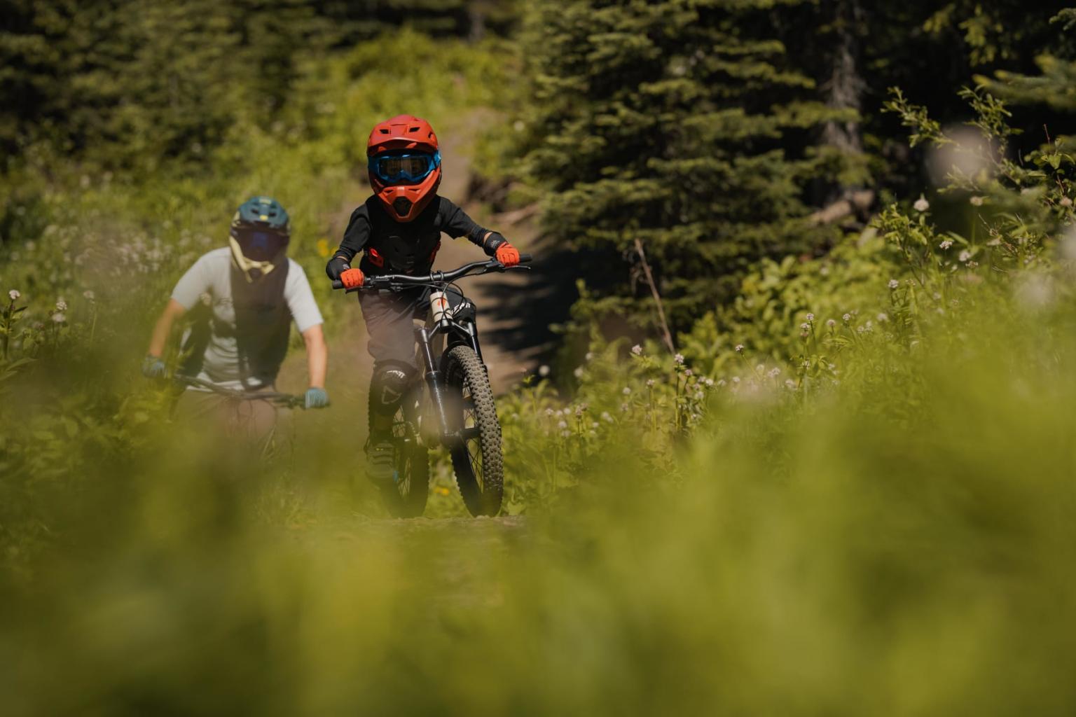 Two cyclists ride through a lush, forested trail wearing helmets.